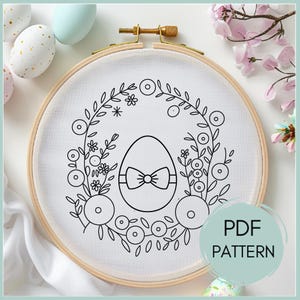 May include: A black and white embroidery pattern of an egg with a bow in a wreath of flowers. The pattern is on a white fabric in a wooden embroidery hoop. The text "PDF PATTERN" is in the lower right corner of the image.