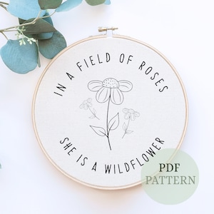 May include: A white embroidery hoop with the text "In a field of roses she is a wildflower" and a black line drawing of a flower with three smaller flowers.