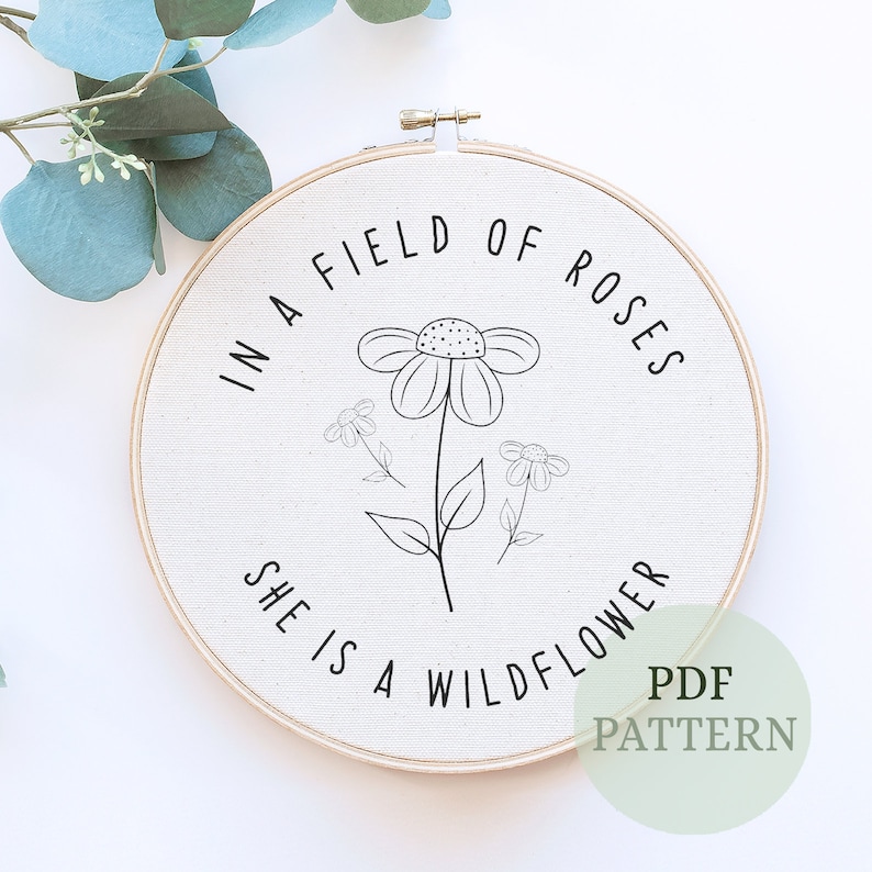 May include: A white embroidery hoop with the text "In a field of roses she is a wildflower" and a black line drawing of a flower with three smaller flowers.