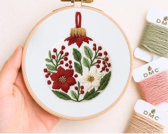 Puede incluir: Un diseño de adorno navideño bordado a mano en un aro de madera. El adorno presenta flores rojas y blancas, hojas verdes y bayas rojas. El texto "Hand Embroidery Template" está en la parte inferior. Se ven carretes de hilo de bordar en verde, rosa y beige.