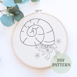 Puede incluir: Patrón de bordado de línea en blanco y negro de una concha con flores. El patrón se muestra en una tela blanca en un aro de bordado de madera. El texto "PDF PATTERN" se muestra en un círculo verde debajo del aro.