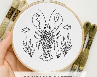 May include: A white embroidery hoop with a black line drawing of a lobster decorated with flowers, two fish, and seaweed. The text "PRINTABLE PATTERN" is at the bottom. Embroidery floss in shades of green and tan are in the background.