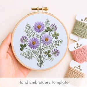 May include: An embroidery hoop with a floral design featuring purple daisies and clover. The embroidery is on a light blue fabric, held in a wooden hoop. Spools of DMC thread in green, pink, and tan are visible. The text "Hand Embroidery Template" is at the bottom.