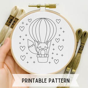 Op de afbeelding: Een witte borduurring met een zwart-wit vos in een luchtballonontwerp. Het ontwerp bevat harten en plustekens. De tekst "PRINTABLE PATTERN" staat onderaan. Borduurgaren is op de achtergrond te zien.