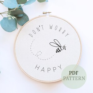 Puede incluir: Un aro de bordado blanco con el texto "No te preocupes, sé feliz" y una ilustración de abeja en blanco y negro. El texto "Patrón PDF" también es visible en la esquina inferior derecha.