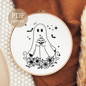 Peut inclure: Motif de broderie noir et blanc d'un fant&ocirc;me tenant une citrouille avec des fleurs et des &eacute;toiles autour. Le motif est sur un fond blanc et se trouve dans un cerceau de broderie rond.