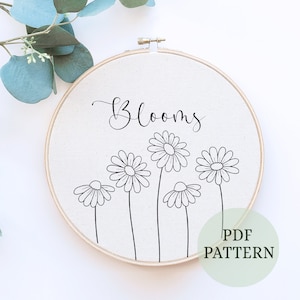 May include: A black and white embroidery pattern featuring the word "Blooms" and five stylized daisies. The pattern is displayed on a white fabric stretched over a wooden embroidery hoop. The text "PDF PATTERN" is written in green on a white background.