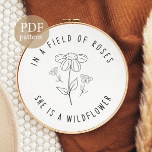 May include: A white embroidery hoop with black embroidery of a flower and the text "In a field of roses she is a wildflower".