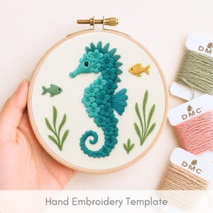 Op de afbeelding: Een borduurring met een turquoise zeepaardontwerp, omringd door gele en groene vissen en zeewier. De tekst "Hand Embroidery Template" staat onderaan. Spoelen garen in groen, roze en beige zijn zichtbaar.