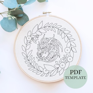 May include: Black and white line art embroidery pattern of a dragon's head surrounded by a wreath of leaves and berries. The pattern is on a white background and is labelled "PDF TEMPLATE".