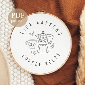 May include: White embroidery hoop with black line art of a coffee pot, coffee cup, and the text "Life Happens Coffee Helps".