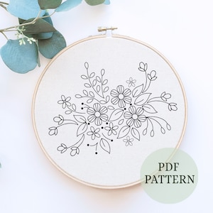 May include: A floral embroidery pattern in a wooden hoop. The design features a cluster of flowers, leaves, and stems in black line art on a white fabric background. The text "PDF PATTERN" is in a green circle.