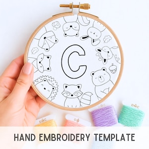 Może przedstawiać: Drewniany tamborek z białą tkaniną z czarnym obrysem litery "C" otoczonej leśnymi zwierzętami. Tekst "HAND EMBROIDERY TEMPLATE" znajduje się na dole. Widoczne są również szpulki kolorowej nici do haftu.