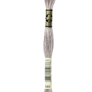 Könnte beinhalten: Ein Knäuel DMC Stickgarn in Farbe 648, ein helles Grau. Das Knäuel ist um eine Pappspule mit einem goldenen Etikett gewickelt, auf dem "DMC" und "25m (82,7 yd)" steht.