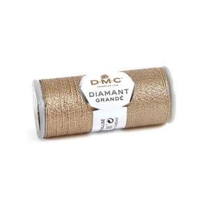 Puede incluir: Un carrete de hilo DMC Diamant Grandé beige oscuro. La etiqueta dice "DMC DIAMANT GRANDÉ". El hilo es de color metálico y brillante. El carrete es cilíndrico con tapas de extremo gris. El texto "G225 - dark beige" está en la parte inferior.