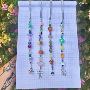 May include: Four vertical beaded chains featuring a variety of colorful plastic and glass beads in shapes including flowers, stars, hearts, and yin-yang symbols. Each chain is finished with a silver-toned metal charm, including a bow and heart.
