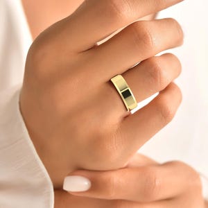 Op de afbeelding: Een goudkleurige ring met een plat, rechthoekig ontwerp. De ring wordt aan een vinger gedragen en toont zijn gepolijste oppervlak. Het sieraad reflecteert licht, wat de metalen glans benadrukt. Een eenvoudig, elegant accessoire.