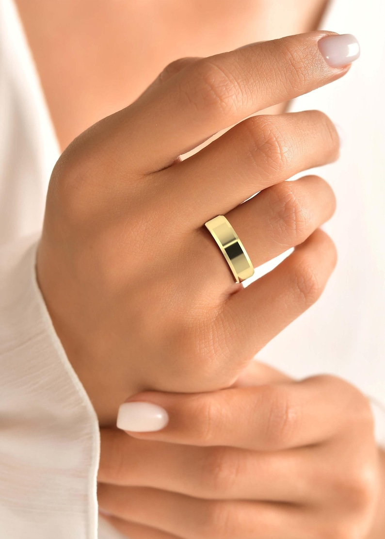 Op de afbeelding: Een goudkleurige ring met een plat, rechthoekig ontwerp. De ring wordt aan een vinger gedragen en toont zijn gepolijste oppervlak. Het sieraad reflecteert licht, wat de metalen glans benadrukt. Een eenvoudig, elegant accessoire.
