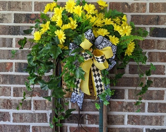 May include: A yellow and black checkered bow with a yellow ribbon hangs from a basket filled with yellow flowers and green leaves. The basket is hanging on a brick wall.