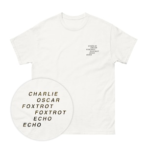 Puede incluir: Camiseta blanca con el texto "CHARLIE OSCAR FOXTROT FOXTROT ECHO ECHO" impreso en dorado en la parte delantera.