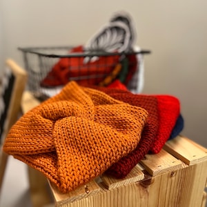 May include: A stack of four knitted headbands in solid colors including burnt orange, dark red, bright red, and navy blue. Each headband features a central twisted knot detail and a thick, textured yarn construction with visible knit stitches.