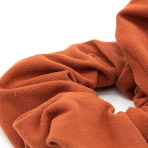 May include: Close-up of a rust-colored scrunchie made of soft fabric. The scrunchie is gathered and has a textured appearance. The background is white, highlighting the details of the hair accessory.