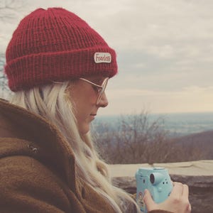 May include: A deep red knit beanie with a white rectangular patch featuring the word Fearless in red script. The beanie has a ribbed texture and a folded cuff. A light blue instant camera is held in the foreground, partially visible in the frame.