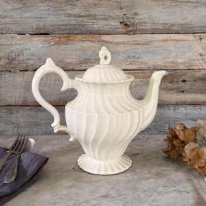 May include: Cream-colored ceramic teapot with a lid and a curved handle. The teapot has a spiral design on the body and a decorative finial on the lid. The teapot is on a marble surface with a fork and spoon on a napkin.