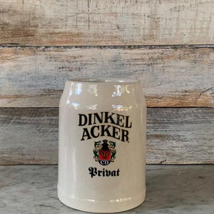 May include: A cream-colored ceramic beer stein with the words "DINKEL ACKER" in black lettering. Below the name is a crest with red and green accents, and the word "Privat" is written below the crest. The stein has a cylindrical shape.