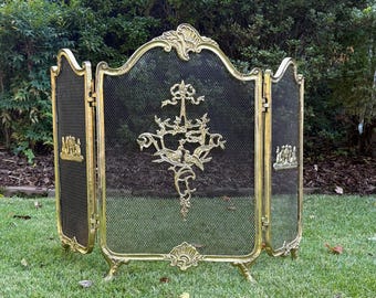 May include: A three-panel brass fireplace screen featuring a fine black mesh center. The central panel displays an ornate gold-toned metal emblem of two birds perched on a branch, while the two side panels feature smaller decorative metal motifs.