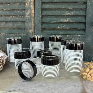May include: A set of eight clear drinking glasses with a black band at the top and a white horizontal striped design. The glasses are cylindrical and sit on a marble surface, with a weathered green shutter background.