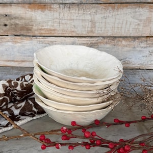 May include: A stack of six off-white, textured bowls with irregular edges. The bowls are stacked on top of each other, with a hint of green visible between some. A brown and white patterned cloth and red berries on a branch are in the foreground.