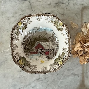 May include: A vintage, octagonal-shaped ceramic bowl with a winter scene illustration. The bowl features a brown rim with floral and leaf patterns. The central image depicts a red covered bridge in a snowy landscape, with trees and a mountain in the background.