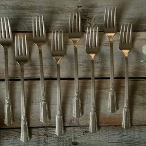 May include: Ten silver forks with ornate handles are arranged in a row on a wooden surface. The forks are all facing the same direction, with the tines pointing upwards.