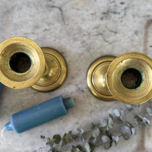 May include: Two brass candlesticks with circular bases and wide rims, viewed from directly above. Two short, cylindrical blue candles are positioned next to the candlesticks, accompanied by a sprig of dried eucalyptus leaves.