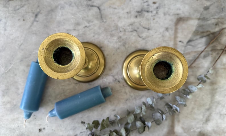 May include: Two brass candlesticks with circular bases and wide rims, viewed from directly above. Two short, cylindrical blue candles are positioned next to the candlesticks, accompanied by a sprig of dried eucalyptus leaves.