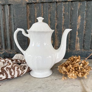May include: A white ceramic coffee pot with a lid, handle, and spout. The pot has a ribbed design and sits on a marble surface. A patterned cloth and dried flowers are also present.
