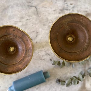 May include: Two circular brass candle holders viewed from above, featuring a weathered, brownish-gold patina. Each holder has a central spike for securing a candle. Two blue pillar candles and a sprig of dried eucalyptus are positioned nearby.