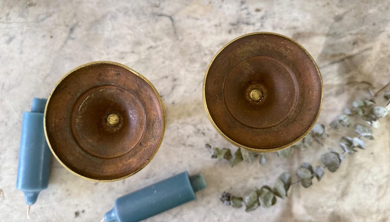 May include: Two circular brass candle holders viewed from above, featuring a weathered, brownish-gold patina. Each holder has a central spike for securing a candle. Two blue pillar candles and a sprig of dried eucalyptus are positioned nearby.