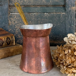 May include: A copper-colored coffee pot with a silver-colored interior and a long, gold-colored handle. The pot has a wide base that tapers to a narrower top. The background includes old books and dried flowers.