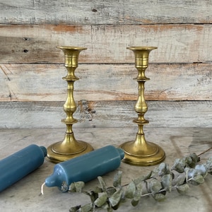 May include: Two matching brass candlesticks with a polished finish, featuring a tiered base and a slender, decorative stem. Beside the candlesticks lie two solid blue pillar candles and a sprig of dried eucalyptus leaves with small green foliage.