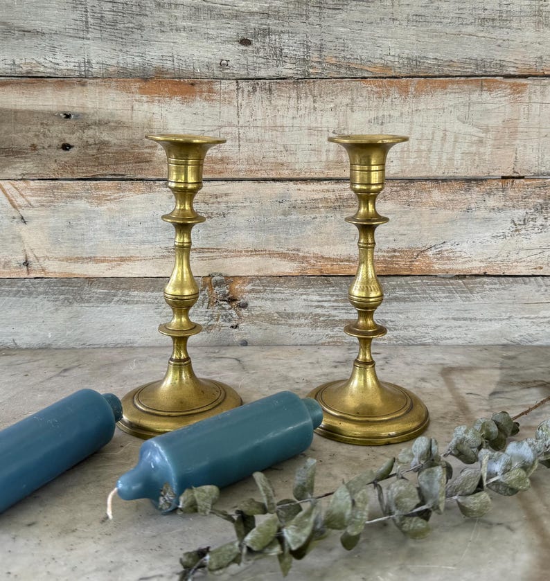 May include: Two matching brass candlesticks with a polished finish, featuring a tiered base and a slender, decorative stem. Beside the candlesticks lie two solid blue pillar candles and a sprig of dried eucalyptus leaves with small green foliage.