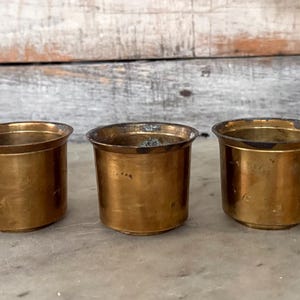 May include: Three small, cylindrical brass containers with a slightly flared rim. The containers have a warm, golden hue and sit on a marble surface. The background is a weathered, light-colored wooden panel.