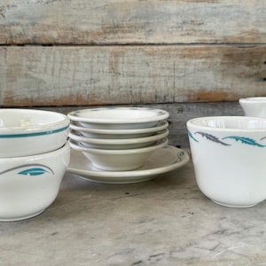 May include: A collection of white ceramic dishware with teal and gray decorative accents. The set includes bowls, saucers, and a mug. The bowls and mug feature a wave-like design. The items are arranged on a light-colored surface.