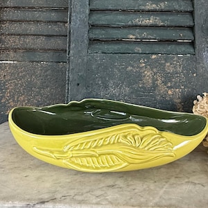 May include: A yellow ceramic planter with a dark green interior. The exterior features a raised leaf design. The planter is oval-shaped and sits on a marble surface. The background includes weathered green shutters.