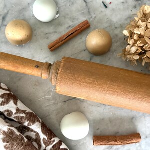 May include: A wooden rolling pin with a light brown finish, alongside cinnamon sticks, decorative balls, and dried flowers. A patterned cloth is also visible. The items are arranged on a marble surface.
