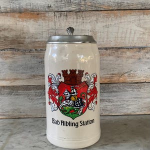 May include: Cream-colored ceramic beer stein with a silver-colored metal lid. The stein features a detailed crest with a knight on horseback and the text "Bad Aibling Station". The crest is framed by red and white decorative elements.