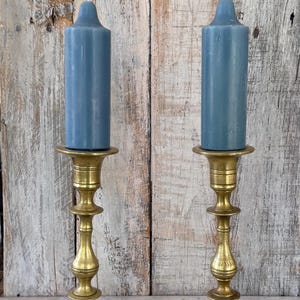 May include: Two matching brass candlesticks with a polished finish and tiered, slender stems, each holding a solid, cylindrical blue candle with a rounded top. The candlesticks feature wide, circular bases and a classic, symmetrical design.