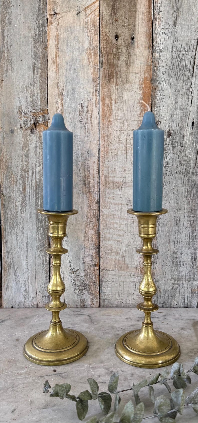 May include: Two matching brass candlesticks with a polished finish and tiered, slender stems, each holding a solid, cylindrical blue candle with a rounded top. The candlesticks feature wide, circular bases and a classic, symmetrical design.