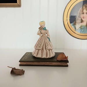 May include: A vintage porcelain figurine of a woman in a long, layered dress, with a light pink and green color scheme. The figurine is placed on a dark green book with gold accents. A dried leaf rests nearby.